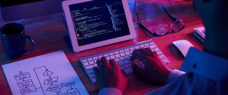 Um programador está sentado à sua mesa, trabalhando à noite. A iluminação de néon azul e magenta do monitor e da sala reflete em sua tela de computador e em seu rosto. Em primeiro plano, o programador está digitando em um teclado branco, com uma xícara azul à esquerda e um fluxograma desenhado à mão em uma folha de papel. No centro da imagem, um tablet exibe código de programação.